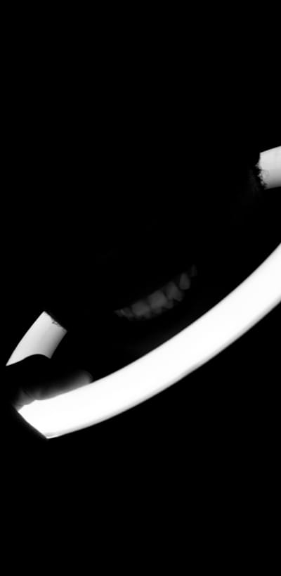 This black and white, high-contrast image presents an abstract close-up, dominated by a bright, curved light source that runs diagonally from the top to the bottom right of the frame. The light appears uniform and intense, suggesting an artificial light source. To the left of this prominent light, a shadowy form reveals several small, lighter, rounded shapes, possibly indicating beaded details on clothing, jewelry, or an object. At the bottom right, a darker, finger-like shape intrudes into the frame, seemingly interacting with or holding the light source. The background is predominantly black, emphasizing the illuminated elements. The scene appears to be taking place in low light conditions, such as at night or in a dark interior. No text is visible.