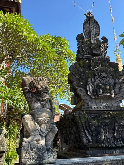 An outdoor, medium shot captures two intricately carved stone statues against a backdrop of lush green foliage and a bright blue sky. The statue on the left depicts a crouching, fierce creature with a gaping mouth, sharp teeth, and ornate headdress. A single pink flower rests on its ear. The statue on the right is a larger, more elaborate structure featuring a winged mythical creature with a dragon-like head and detailed carvings of other figures. The environment suggests a temple or traditional Balinese courtyard, possibly in Ubud District, Indonesia, given the style of the carvings and the presence of a distant yellow and green ceremonial banner. The lighting indicates a sunny day.