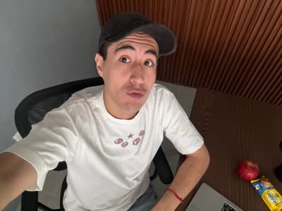 A young man is captured in a selfie-style shot, seated at a desk in what appears to be a home office or study. He is wearing a white t-shirt with a graphic design featuring dice and a star, and a dark baseball cap. His facial expression is a playful pout. To his right, on the wooden desk, are an apple and a packaged snack. The background shows a textured wooden wall and a grey wall. The room is dimly lit, with the primary light source appearing to be from above and to the left of the subject. The image does not provide any specific visual cues to pinpoint a location in Puebla, Mexico.