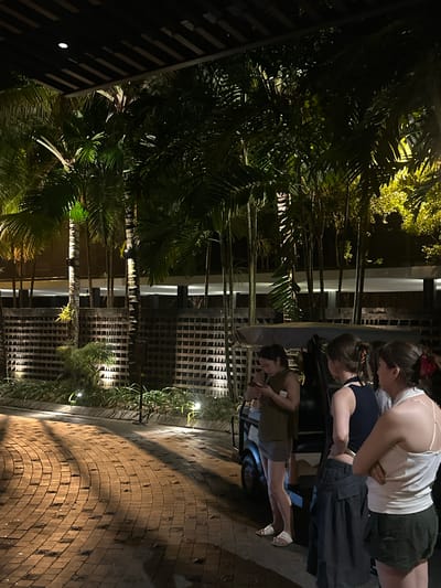 This image depicts a nighttime scene in Kuta, Indonesia, featuring a group of people gathered around what appears to be a golf cart. The setting is outdoors, with lush palm trees and tropical foliage evident, illuminated by artificial lighting. In the background, a building with a distinctive latticed facade is visible. The ground is paved with brickwork, suggesting an outdoor entertainment area or walkway.
There are three individuals clearly visible. Two women are leaning over and interacting with something inside or near the golf cart. One of them is wearing a white tank top and dark shorts, while the other has on a dark top and dark pants or a skirt. A third person, also appearing to be a woman, is standing to the side of the golf cart, looking down. The lighting suggests it is nighttime, and the ambient illumination casts shadows and highlights on the scene. The overall mood seems casual and social, with the individuals engaged in conversation or an activity.
Notable details include the tropical vegetation and the unique latticed architectural element, which could be a design feature of the establishment. The presence of the golf cart suggests a resort, a golf course, or a similar recreational venue. No visible text is present in the image. The focus is on the interaction between the individuals and their surroundings at night.