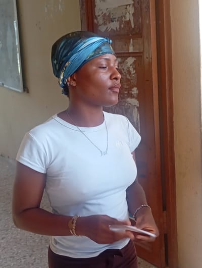 A woman stands indoors, possibly in Ukwu-Abwa, Nigeria, although the location cannot be definitively confirmed from the image alone. She wears a blue headscarf, a white t-shirt with the word "alo" printed on it, a silver necklace, and a brown skirt. She also has jewelry on her wrists. She holds a white object in her hands. Behind her is a door with peeling paint and an off-white wall. A board is visible on the wall to the left. The floor appears to be tiled.