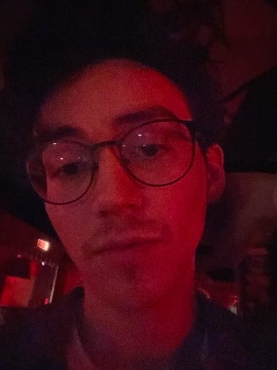 The image presents a close-up portrait of a single individual, identified as male, illuminated predominantly by strong red ambient lighting within an indoor commercial establishment. The subject occupies the majority of the frame, facing directly towards the camera with a neutral expression. He wears dark, circular-rimmed eyeglasses and has short, dark hair. A faint mustache is visible. The upper portion of a dark garment is discernible around his neck and shoulders. The background is dark and out of focus, with a rectangular light source, possibly an emergency exit sign, faintly visible to the left of the subject. The overall scene is dimly lit and characterized by significant red light saturation. No specific identifying features are present to precisely determine the location within the city of Bogota, Capital District, Colombia, beyond it being an interior commercial space such as a bar or nightclub.