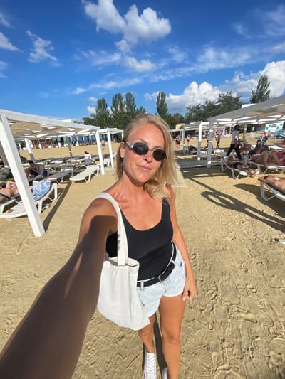In a wide shot taken on a sandy beach, a woman with blonde hair and sunglasses stands facing the camera, holding a white tote bag. She is wearing a black tank top, light-wash denim shorts, a black belt, and white sneakers. The beach is lined with white cabanas and lounge chairs. In the background, other beachgoers are visible, some relaxing on chairs and others walking. Trees and a blue sky with scattered clouds form the backdrop. The overall scene suggests a sunny day at a resort. The location is the beach in Anapa, Russia.