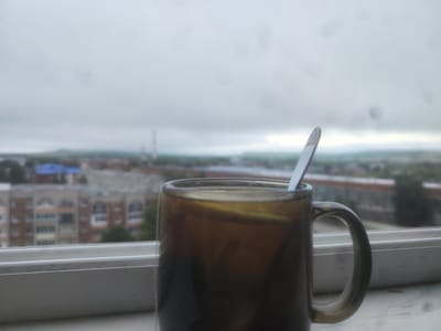 A mug of brown liquid with a spoon is positioned on a windowsill, overlooking a blurry urban landscape. The city of Vyatskiye Polyany, Russia, is visible in the background, with buildings and trees appearing indistinct due to the shallow depth of field. The sky is overcast, suggesting a cloudy day. Water droplets are present on the windowpane, adding to the overall soft focus of the scene.
