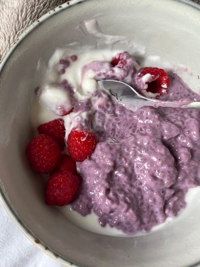 A close-up overhead shot shows a bowl of chia pudding mixed with yogurt and raspberries. The pudding is a vibrant purple color, with small dark specks that are the chia seeds. Swirls of white yogurt are mixed throughout, and several bright red raspberries are visible, some whole and some partially submerged in the pudding. A silver spoon is partially visible in the upper right portion of the bowl, with its tip resting in the pudding. The bowl itself is a neutral, speckled ceramic color, with a slightly textured rim. The background consists of a textured, beige fabric, possibly a placemat or towel, and a white surface, possibly a tablecloth. The image captures a healthy and visually appealing breakfast or snack. There is no indication of the city of Aarhus, Denmark, or any specific location.