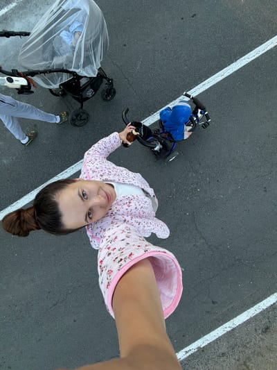 This is an overhead, wide-angle, slightly tilted shot on asphalt, likely taken outdoors in Красный Маяк, Russia. The central subject is a young woman with dark hair pulled back in a bun and wearing a floral-patterned blouse and a white shirt. She is holding a bottle in her right hand and looking directly at the camera with a slight smile, appearing to take a selfie.
To her left is a child's blue tricycle with a blue garment on the seat, positioned next to a white line painted on the asphalt. Further to the left, a black stroller covered with a clear plastic rain cover is visible, with a hint of a baby or child inside. Another person, likely an adult, is standing in the lower left of the frame, wearing light gray pants and green shoes, with their legs and feet partially visible.
The scene suggests a leisurely outing, possibly a walk or a trip to a park. The lighting appears to be natural daylight, suggesting it is daytime, but the absence of strong shadows might indicate overcast weather or late afternoon. The asphalt surface and the presence of painted lines point to a road, a sidewalk, or a paved recreational area. The woman's expression is relaxed and engaging, implying a pleasant and casual moment. No prominent text is visible in the image.