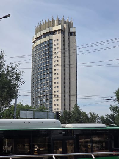 This media file depicts a tall, rectangular building with many windows, likely an apartment building or office block, set against a clear, pale blue sky. The building has a light-colored facade, possibly brick or stone, and features a distinctive, stepped roofline with what appear to be decorative elements. Numerous thin, dark cables crisscross the frame in front of the building, suggesting the presence of overhead power lines or telecommunications infrastructure.

To the right of the frame, a portion of a modern, green and white vehicle is visible. Its dark, reflective windows reveal the interior, showing some seating and structures. This vehicle appears to be a public transport bus or tram. Trees with green foliage are scattered throughout the scene, framing the building and the vehicle, and suggesting an urban or suburban setting.

The perspective is from a low angle, looking up at the building and the sky. The lighting suggests it is daytime, likely morning or afternoon, with bright, diffused sunlight. The overall mood is neutral, conveying an urban landscape with everyday infrastructure. No people are clearly discernible, and no visible text is present in the image. The scene is consistent with an urban environment in Almaty, Kazakhstan, as indicated by the context.