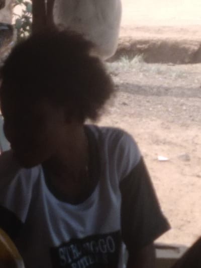 The image displays a close-up of an individual located in Agege, Nigeria, viewed from the chest up. The person has dark, curly hair, with their face obscured by shadow and blur. They are wearing a short-sleeved t-shirt; the main body is light-colored, and the sleeves are dark. The word "STRONGGO" is partially visible in dark lettering on the front of the shirt. A thin, light-colored chain or necklace is present around the individual's neck. The person is looking downwards and towards the left of the frame. The background consists of reddish-brown dirt ground with sparse patches of green vegetation. An indistinct, large, light-colored, rounded object is visible in the upper right background, and a dark, vertical element is partially visible on the upper left, partially obscured by foliage.