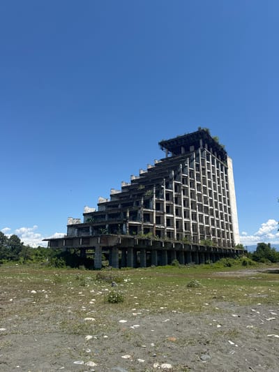 The image captures a large, derelict multi-story concrete building under a clear blue sky in Warcha, Abkhazia. The structure, which appears to be a former hotel or resort, exhibits a distinctive stepped or terraced architectural design, particularly on its left side, where upper floors progressively recede. Its facade is a grid of empty window openings, revealing the exposed concrete framework. Signs of abandonment are prevalent, with green vines and small plants growing on ledges and within the structure itself.

The foreground and the area to the right of the building consist of a mix of dry, sparse grass and darker earth or sand. Numerous scattered, light-colored objects, resembling seashells or weathered stones, are visible across this ground, suggesting a proximity to a natural body of water like a sea or river.

In the background, a line of green trees and distant hills or mountains can be seen beneath the horizon. The sky is a vibrant clear blue with a few fluffy white clouds visible in the upper right. The scene is bathed in bright daylight, indicating midday or early afternoon conditions. No people are present, and no visible text can be deciphered. The image conveys a strong sense of decay, abandonment, and nature gradually reclaiming the man-made environment.