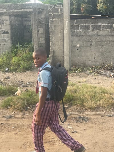 A young Black male, approximately 10-14 years old, walks from right to left on an unpaved street in Mosafejaw, Nigeria. He wears a light blue short-sleeved collared shirt with red and white trim, a red and white striped tie, and red and white plaid trousers. A black backpack with red accents is worn on his back. He looks back over his right shoulder towards the viewer with an open-mouthed expression.
The immediate ground is a dirt path with patches of dry grass. To the boy's left, a light-colored chicken is partially visible foraging in the grass. In the background, a weathered grey concrete block wall stretches across the frame. This wall features two vertical openings, suggesting a dilapidated gateway or incomplete structure. Green overgrown vegetation is visible behind and above the wall. To the right, a grey utility pole stands vertically. The wall shows multiple small circular indentations. Scattered debris and refuse are present on the dirt ground. The scene is brightly illuminated by daylight.