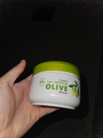 A light-skinned human hand holds a white, cylindrical plastic cosmetic jar with a lime green screw-top lid. The jar's label features green text on a white background, reading "ORIGEN 97% NATURAL" on the upper left, "Cream con ACEITE de OLIVE" prominently in the center, and "deliplus" below "OLIVE". A graphic depicting a cluster of green olives on a small branch is printed on the right side of the label. The hand positions the jar with its base resting on the palm, presenting the label clearly to the viewer. The background is uniformly dark and featureless, appearing black. No environmental details are present to determine a specific location, including any city like Fez, Morocco.