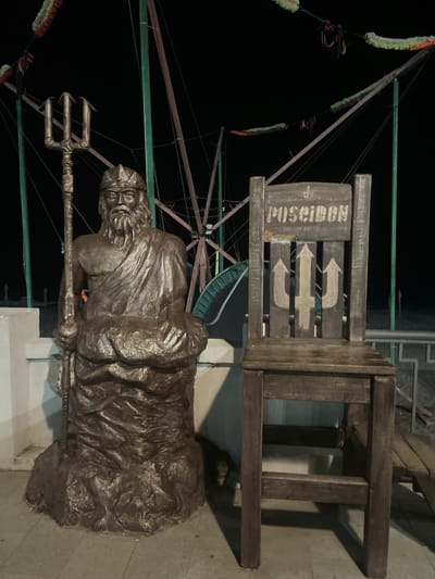 The image shows a bronze statue of Poseidon holding a trident next to a wooden chair with the word "POSEIDON" inscribed on its backrest and a trident symbol painted below. The statue and chair are positioned on a paved area, possibly a walkway or plaza. In the background, there are structures that appear to be part of a carnival or amusement park, with colorful decorations strung overhead against a dark night sky. The setting is identified as Gagra, Abkhazia, suggesting it is a public display or attraction in the city. The lighting is artificial and casts shadows, indicating it is nighttime. There are no people visible in the frame.
