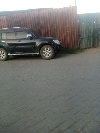 The image displays a black, four-door sport utility vehicle (SUV) parked facing right on a dirt shoulder alongside a corrugated metal fence in Mtopanga ward, Kenya. The SUV exhibits silver roof rails, chrome accents on the door handles and side mirrors, and a running board. The front tire visible on the vehicle's right side is an all-terrain type with "ROADCRUZA" lettering visible. The fence, primarily composed of vertically oriented reddish-brown corrugated metal sheets, transitions to a section of blue-green corrugated metal on the right. Sparse green vegetation is visible at the base of the fence, behind the SUV's front wheel. The foreground and bottom half of the scene feature a paved surface constructed from dark grey rectangular interlocking pavers arranged in a herringbone pattern. The sky above is bright and diffuse. No individuals are present in the scene.