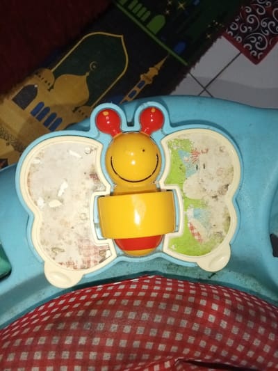 This image captures a light blue plastic children's activity toy located in Ciputri, Indonesia. The toy features a central yellow insect-like figure with red spherical antennae and a red lower body, displaying a simple smiling face. This figure is integrated within a larger yellow cylindrical component, suggesting it is a rotating or spinning element. On either side of the central figure are two white-framed sections resembling wings. The left section contains heavily worn and faded imagery. The right section displays a partially visible and faded cartoon-style illustration, likely depicting an animal against a green background, with a small red and white checkered pattern visible near its base. The toy rests on a red and white checkered fabric surface, which appears to be part of a cushion or seating. In the upper background, a dark-colored prayer mat, adorned with gold and blue mosque and minaret architectural patterns, is partially visible on what appears to be a floor.