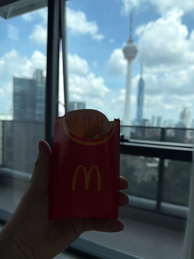 In the foreground, a hand holds a red McDonald's french fry container, its iconic golden arches logo visible. The container is partially filled with french fries. The background depicts a cityscape through a window, with tall buildings and a prominent tower, identifiable as the Kuala Lumpur Tower (Menara Kuala Lumpur), piercing the sky. The sky is blue with scattered white clouds. The scene suggests an indoor perspective looking out over the urban landscape of Kuala Lumpur, Malaysia.