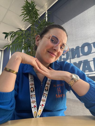 The image shows a female individual indoors, likely within Clinique Al Hayat in Meknes, Morocco. She is seated, facing the camera, with a slight smile. She has dark hair, pulled back, and wears light-colored, hexagonal-rimmed glasses. Her attire includes a blue fleece jacket with a zipper closure. Embroidered on the left side of the jacket is "AL HAYAT R Mery." Around her neck hangs a white lanyard featuring cartoon nurse figures, hearts, stethoscopes, and text such as "KEEP CALM I'M A NURSE." On her left wrist, she wears a gold-toned bangle bracelet, and on her right wrist, a gold-toned metal link watch. Her hands are positioned together in front of her chest, with fingers slightly interwoven. The background includes a tall artificial plant with green leaves and white beaded decorations on the left. To the right, a textured window blind is visible, displaying a faint, reversed shadow-reflection of text. The ceiling features white rectangular tiles, and a light-colored surface is present in the foreground.