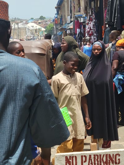 In Suleja, Nigeria, a street scene features a vendor selling clothing and other wares. A young boy in a light brown shirt stands smiling, positioned between women in headscarves, with one woman wearing a black niqab. A man in a blue patterned shirt is present in the foreground. In the background, there is a street scene visible including buildings and a tricycle. A "NO PARKING" sign is partially visible on the right side.