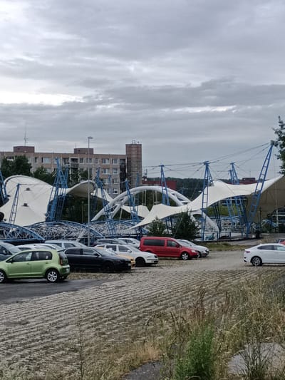 A cloudy day with overcast skies hangs over a parking lot in Prague, Czechia. In the foreground, a textured ground cover, possibly pavement or compacted earth with a grid pattern, stretches across the scene, with dry grasses and weeds growing along the right edge.
A row of parked cars occupies the middle ground. Among them are a bright green hatchback, a black sedan, a white car, and a red van. Behind the cars, a large, modern, tent-like structure with a white fabric canopy is supported by a complex framework of blue metal poles and beams. The canopy appears partially collapsed or in a state of disrepair, with some sections sagging and others dramatically contorted. The structure's intricate design, with its curves and diagonal supports, suggests it might be a temporary venue or pavilion.
In the background, a multi-story building with numerous windows and a reddish-brown facade stands prominently. Smaller structures and trees are visible further in the distance, as are parts of what appear to be other urban elements. The overall atmosphere is somber, possibly due to the weather and the damaged state of the tent structure. No text is visible in the image.