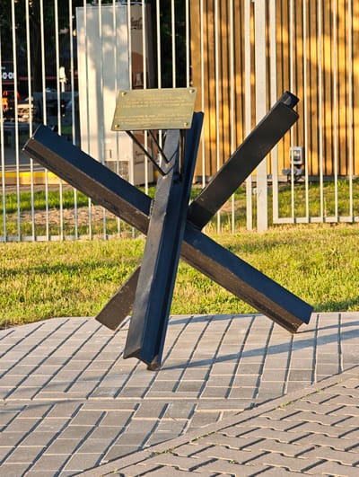 The image shows a monument consisting of a black metal anti-tank obstacle, resembling a three-pronged structure, erected on a paved area. A plaque is affixed to a support behind the obstacle. The background features a metal fence and some greenery. Based on the text on the plaque, this monument is located in Brest, Belarus, and is dedicated to the memory of railway workers and in honor of the 80th anniversary of the Victory of the Soviet people in the Great Patriotic War. The monument is situated outdoors on a sunny day, with long shadows cast on the pavement. No people are visible in the image.