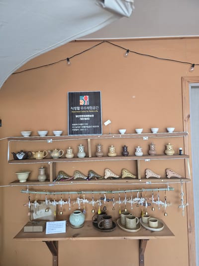 This media file captures a close-up view of a display of ceramic crafts, likely an exhibit at a rural experience center in Gumi-si, South Korea. The display features a variety of handcrafted ceramic items, including ornate whistles resembling ocarinas, small teapots and cups, and decorative pieces. The items are arranged on wooden shelves and racks, with some hanging from thin wires adorned with small, fish-like charms and bells, suggesting a playful and artistic presentation.

The background is a warm, light brown wall, with a portion of a window frame visible at the top. A string of small, exposed light bulbs runs vertically along the wall to the left, hinting at a decorative lighting element. To the left, a large, draped white fabric partially obscures the view, possibly part of a tent or display structure.

A prominent sign in Korean, featuring colorful graphics, is mounted on the wall to the left of the ceramic display. The text on the sign translates to "Experience Center for Dietary Life," and indicates that this is the "Gungsan Eco-friendly Experience Farm (Gungsan Shelter)," designated as an excellent rural experience space by the Ministry of Agriculture, Food and Rural Affairs. It also mentions a date, "2021. 6. 7," likely the designation date. Another smaller sign with Korean text is visible within the display area, providing details about the pottery, mentioning it is handmade and expressing a wish for good fortune, with a price and bank account information.

The overall impression is one of a well-curated exhibition showcasing traditional Korean craftsmanship and promoting a connection to rural life and dietary experiences. The lighting and arrangement of the ceramics create an inviting and perhaps educational atmosphere. There are no people visible in the frame.
