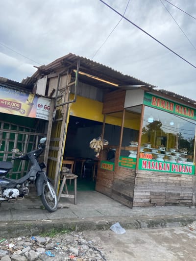 The image shows a small restaurant with a sign that reads "Rumah Makan Masakan Padang," indicating it serves Padang cuisine. The restaurant is located in Katapang, Indonesia, at coordinates -6.993 latitude and 107.556 longitude. The building has a wooden frame with a yellow painted wall and a green awning. The interior of the restaurant is visible through a large glass window, which shows wooden tables and chairs, as well as a variety of dishes on display. A black motorcycle is parked in front of the restaurant. A small cat is sitting on a stool outside the restaurant. There is debris on the ground in front of the restaurant, including a plastic bag and pieces of broken brick. The sky is overcast.