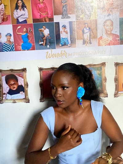 A young Nigerian woman is posing in front of a wall decorated with framed photos and a large banner. The banner displays the text "Wonder Shot PHOTOGRAPHY" in a decorative font. The framed photos appear to be portraits and studio shots, some featuring children in various outfits and poses. The woman is wearing a light blue dress and a large, vibrant blue flower earring with dangling beads. She is also wearing a gold bracelet. Her hair is pulled back into a ponytail. The background wall is white. The lighting suggests an indoor setting, likely a photography studio or a home decorated with studio backdrops. The provided context about the location in "Gwong, Nigeria" indicates the potential cultural and geographical setting of the scene.