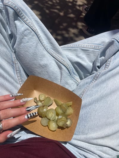 A high-angle shot shows a person seated, wearing light-wash denim jeans, with a small brown cardboard tray of sliced pickles resting on their lap. The person's hand is visible on the tray, showcasing long, pointed nails with various black and white designs, including stripes, a Union Jack, and a dollar sign. The person is also wearing at least two rings. To the left, part of a black shoe can be seen. To the right of the tray is part of a burgundy-colored shirt. The setting is outdoors with harsh sunlight. There is no evidence to confirm the location in the city of Raanana, Israel.