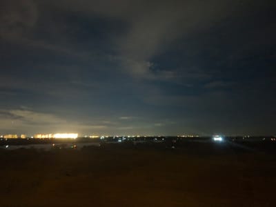 The media file depicts a nighttime cityscape from an elevated viewpoint. The foreground is a dark, textured expanse, possibly barren land or a rooftop. In the mid-ground, a dark body of water or low-lying terrain separates the foreground from the distant city lights.
The background is dominated by a city illuminated at night. A long stretch of buildings is visible, many with bright, warm yellow lights, suggesting residential or commercial structures. Scattered among these are smaller, brighter white lights, which could be streetlights, individual buildings, or vehicles. The sky above is dark, with hints of clouds that are faintly illuminated by the city glow. The overall impression is of a sprawling urban area at night, seen from a distance.
The time of day is clearly night, and the weather appears to be clear with some cloud cover, as evidenced by the visible lights and the dark sky. There are no discernible people or specific objects in the foreground or mid-ground, the focus being on the distant urban landscape. The scene evokes a sense of quiet observation of the city from afar. No text is visible in the image.