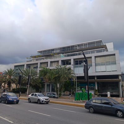 The image depicts a cloudy day in Caracas, Venezuela, with a multi-story modern office building as the primary subject. The building features a glass facade, balconies with glass railings, and several levels, including a rooftop with greenery and seating areas. Palm trees and other tropical vegetation are visible in front of and around the building.
In the foreground, a two-lane road runs horizontally across the frame. Several cars are present on the road: a blue hatchback is in the left lane, and a silver sedan is in the right lane. Further down the road, a dark-colored sedan is in the right lane, captured in motion. A person is seen walking on the sidewalk in front of the building, near a large green sign that appears to spell out "ATURIA". The building's ground floor has visible signage for "BANCO CARIBE". The overall scene suggests an urban commercial district with contemporary architecture and a tropical climate.