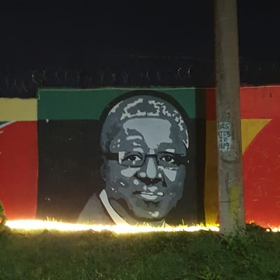 The image shows a mural of a man's face on a wall, likely in Barakin Lerawi, Nigeria. The mural is rendered in shades of gray and white, with the subject wearing glasses and having short, wavy hair. The background of the mural features horizontal bands of color: yellow, green, and black. To the right of the mural, a concrete pole is visible, with white graffiti on it that appears to read "NES AJSS JB 0119". The wall is bordered at the top by barbed wire. In front of the wall, there is a strip of bright, yellowish light, possibly from streetlights, illuminating the grass and the lower part of the mural. The overall scene is dimly lit, suggesting it was taken at night.