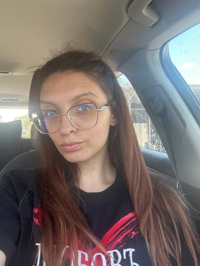 A young woman with long, reddish-brown hair and blue eyes is seated in the driver's seat of a car. She is wearing large, square-shaped glasses with thin gold frames and a black t-shirt with a red graphic design and white text. Her hair is partially pulled back from her face, and some strands fall loose around her shoulders. The interior of the car is visible, including the headliner, a grab handle, and the door frame. Through the car window, a blurred background of trees and a building can be seen. The sky is a clear blue. The image appears to have been taken during the daytime, with natural light illuminating the scene. The location is not specified in the image itself, and there is no information available to suggest it is in the city of Tryavna, Bulgaria.