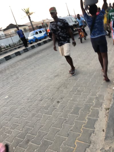 The image depicts a street scene in Moba, Nigeria. The ground is paved with interlocking gray bricks. Two individuals are visible, seemingly walking on the pavement. One is wearing a dark patterned shirt and light-colored shorts, while the other is wearing a blue patterned shirt and dark shorts. A blue and white vehicle, possibly a bus, is parked on the side of the street. There are trees in the background, and buildings are visible in the distance. A metal fence separates the pavement from the street.