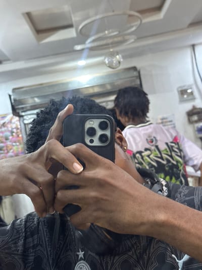 A person holds a smartphone to take a selfie in a mirror. The reflection in the mirror shows another person wearing a white jersey with black and green patterns. The location is Ogwashi-Uku, Nigeria, based on the prompt. In the background, there's a fan and a partially visible window. The lighting appears to be indoor lighting. The person holding the phone is wearing clothing with patterns.