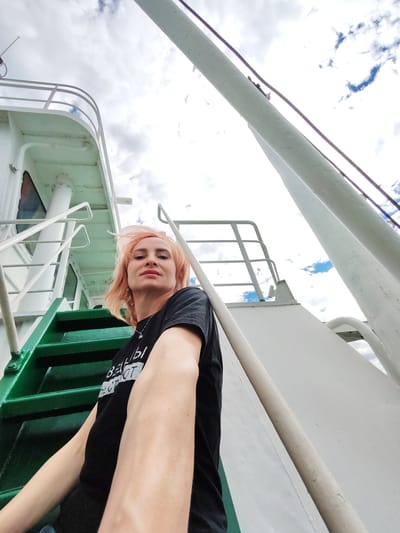 A woman with light-colored hair and wearing a black t-shirt with white text is posing for a selfie on the deck of a white ship. She is positioned on green metal stairs, looking towards the camera. The background shows the upper structure of the ship, including white railings and large white pipes, against a sky with white clouds and patches of blue. The woman appears to be in the port of Шебунино, Russia, given the context.
