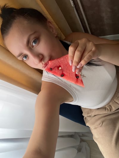 In this media file, a young woman is captured in a close-up shot, holding a slice of watermelon up to her face. Her eyes, a striking blue, are wide and looking directly at the camera, conveying a sense of playfulness or curiosity. She is wearing a white ribbed tank top and a beige corduroy skirt. The watermelon slice is positioned so that a part of it obscures her mouth, and her fingers, with neatly manicured nails, are gently holding it in place. A small silver cross earring is visible on her ear. The background suggests an indoor setting, with a warm-toned curtain to her left and sheer white curtains to her right, hinting at a window. The lighting appears soft and natural, possibly indicating daytime. The overall mood is casual and intimate. There is no visible text in the image.