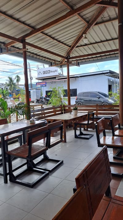 The image depicts an outdoor seating area of a cafe or restaurant, likely in Julikeude Dua, Indonesia, given the signage. The area is covered by a corrugated metal roof supported by wooden beams and posts. Several wooden tables with matching benches and chairs are arranged on a tiled floor. In the background, a white building with a prominent sign that reads "EDK Kuphi" is visible, along with other structures and some vegetation. A dark-colored car is parked in front of the building, and two motorcycles are parked to the left. The scene appears to be during daylight hours with overcast skies.