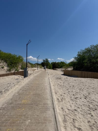 The image captures a sunny outdoor scene in Liepaja, Latvia, depicting a wooden boardwalk path cutting through a sandy dune environment. The path, composed of horizontal wooden planks, extends from the foreground into the distance, curving slightly to the right. Faint yellow markings are visible on the boardwalk planks in the foreground. On both sides of the path, light-colored sand dunes rise, populated with green bushes and scattered small trees. A segmented, low wooden fence constructed from vertical logs runs along the base of the dune on the left side of the path, and a similar fence segment is visible further down on the right. Tall, black, modern streetlights with distinct angular tops are installed at intervals along the left side of the boardwalk; a dark gray utility box is positioned at the base of the nearest streetlight. An adult individual, viewed from behind, wearing a white shirt and light-colored shorts, is walking away from the camera along the boardwalk, carrying a dark bag in their left hand. The sky is clear and bright blue, with a few small white cumulus clouds visible above the distant dunes. Footprints are discernible in the sand bordering the boardwalk.