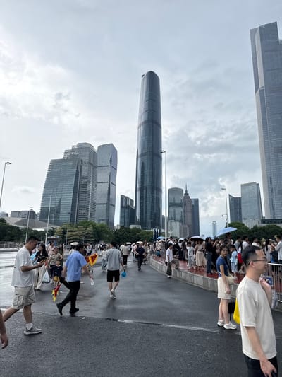 This image captures a street scene in Tianhe District, China, on a cloudy day. A large crowd of people is gathered on a wet asphalt surface, which appears to be a pedestrian walkway or street. In the background, a cluster of modern skyscrapers dominates the skyline. The architecture of these buildings is characterized by glass facades and varied geometric shapes. One particularly tall, cylindrical building stands out, while others are more rectangular.

The people in the foreground and midground are walking and standing, some holding umbrellas. Their clothing suggests a casual setting, with many wearing t-shirts and shorts. A few individuals are holding small, colorful flags or banners. The ground is wet, indicating recent rain or a damp environment. The overall atmosphere is one of public gathering, possibly for an event or simply navigating the urban space. The sky is overcast with diffused light, contributing to a muted color palette.