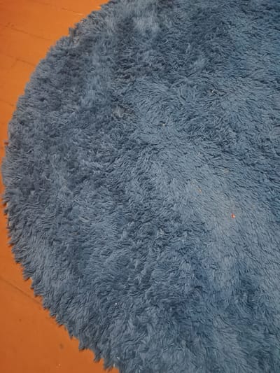 This is a close-up, overhead shot of a circular, fluffy, dark blue rug lying on a wooden floor. The rug has a shaggy texture, with long, soft fibers creating a plush surface. The wooden floorboards are a warm, orangey-brown color and appear to be old, with visible grain and some subtle variations in tone. The rug covers most of the frame, with its edge visible in the lower left corner. The lighting is somewhat dim, suggesting an indoor setting, possibly at night or in a room with limited natural light. There are a few tiny specks of color, a red and some light brown, visible on the rug, which could be dust or small debris. The overall impression is one of a cozy, domestic environment. No people are visible in the image. The location context of Donetsk, Ukraine, does not provide specific visual cues within the image itself, but it places the scene within a particular geographical and geopolitical setting. There is no visible text.