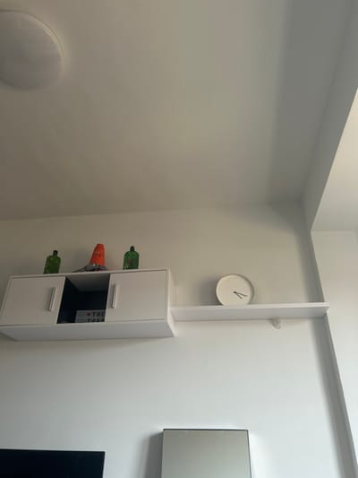 The image shows a white wall with a white cabinet, a white shelf, a white clock, and a mirror. The cabinet has two white doors, and one black door. The clock is round with black hands. The cabinet has a small red light inside it that reads "THE TRAP" on a white background. The shelf is attached to the wall above the cabinet. There is a green glass bottle and a green glass jar sitting on the cabinet. The mirror is attached to the wall at the bottom of the image. The scene is located in Segovia, Spain, at the coordinates 40.951 latitude and -4.121 longitude. There are no people present.