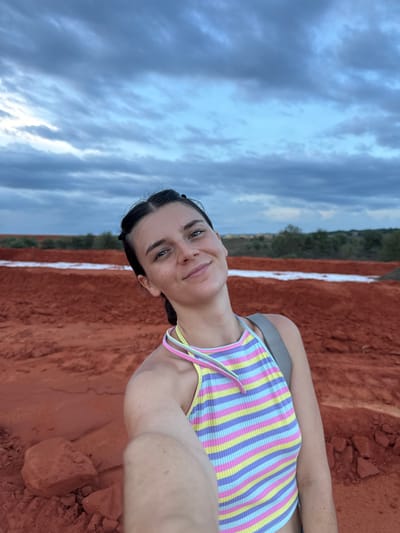 A young woman with dark hair pulled back into two small buns on top of her head, and freckles across her nose and cheeks, smiles at the camera in a selfie. She is wearing a multi-colored striped tank top with thin straps in yellow, pink, purple, and blue. She is also wearing a grey backpack strap over her right shoulder.
The background appears to be a rural or industrial landscape, with large mounds of reddish-orange dirt. A thin strip of white material runs horizontally across the middle ground. In the distance, there are low green trees and hills under a cloudy, blue-grey sky. The overall impression is of an outdoor, possibly arid or construction site, environment. This location is likely the sand dunes of Mui Ne, Vietnam, known for their distinct red sand.