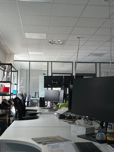 This image captures a modern, brightly lit office environment, likely a shared workspace, viewed from a slightly elevated perspective looking across multiple desks. The scene appears to be devoid of people, suggesting either an early morning, late evening, or unoccupied workspace.

In the foreground and to the right, a desk is visible, featuring several computer monitors, mostly black, with one showing the brand name "iiyama" and another partially revealing "G-MASTER." A black keyboard and mouse are present, alongside a white tissue box with a subtle green leaf pattern. A small blue plush toy, resembling a character with prominent eyes, sits on the desk next to a striped object that partially reads "LOLA." Various papers and organizers are scattered across the desk, indicating a working environment.

The midground reveals a series of office chairs, predominantly black with mesh backs, positioned at other desks. Laptops, including a silver HP model, and other office supplies are also visible on these workstations. The space is structured by sleek glass partitions with dark frames, dividing different work areas and creating an open yet organized feel.

Towards the left and further back, the architectural elements of the room become more prominent. The ceiling consists of white square acoustic tiles, interspersed with circular light fixtures or vents. A thin cable runs horizontally across the ceiling. On the bottom right of the image (which would be the floor level if the image were correctly oriented), shelving units are visible, holding files and various items, including one item with "walton" partially legible.

The overall impression is one of a clean, functional, and contemporary office, ready for productivity. The lighting appears artificial, suggesting an indoor setting independent of direct natural light, though the general brightness implies daytime.