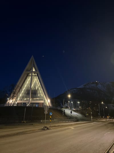The image displays a nocturnal scene in Tromsø, Norway, featuring a distinctive modern building, a road, and a snow-covered mountain under a dark blue sky. The prominent subject in the mid-ground is a building with a striking triangular or A-frame glass facade, brightly illuminated from within, revealing its internal structure and multiple levels. Bare tree branches are visible in front of the building.

To the right of the building, an asphalt road extends into the distance, appearing wet or possibly icy. White and yellow lane markings are visible on the road surface. Multiple streetlights cast a warm, yellow glow, illuminating the road and parts of the surrounding environment. Several traffic signs are present along the roadside, including a blue circular sign with a white arrow and a white circular prohibitory sign with a red border and diagonal line, though no text is legible.

In the background, a large, dark mountain or hill, partially covered in snow, rises steeply against the night sky. Scattered lights can be seen on its slopes, suggesting structures or residences. The overall impression is one of a cold, clear winter night in a northern city, with artificial light creating a stark contrast against the dark natural landscape. No people are visible, and no specific events or activities are overtly occurring.