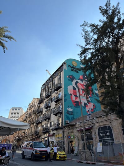 An outdoor, daytime, eye-level shot captures a street scene in Jerusalem, Israel. The dominant feature is a large building with a vibrant teal mural depicting a stylized bird. The words "PARAMOUNT HOTEL" are visible in gold lettering above the mural. To the right, a large pine tree partially obscures the view of another building. In the foreground, an ambulance with red and white markings is parked on the street, with two men standing near it. A yellow taxi is also present. Several pedestrians are visible walking on the sidewalk. The street is lined with multi-story buildings, some with balconies. A construction crane is visible in the distance. The sky is clear and blue.