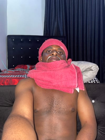 An indoor scene, identified as being in Ube, Nigeria, features a single adult male, photographed from the chest up. The man has dark skin and visible chest hair, and is shirtless. He wears a red terry cloth towel wrapped around his head like a turban, and another red terry cloth towel draped around his neck and shoulders. His face exhibits white markings: a thin line above his upper lip resembling a mustache, and several small dots on his cheeks and forehead. He is looking directly at the camera, holding the device with his right arm, which is visible in the lower right foreground. The background reveals a bedroom setting, including a dark, tufted headboard with decorative buttons on the left, positioned against a light wall. A bed with a red and black patterned duvet or sheet is partially visible below the headboard. To the right, dark curtains are present, alongside a light-colored blanket or pillow and a black and white patterned item on the bed. The scene is illuminated by what appears to be artificial light.