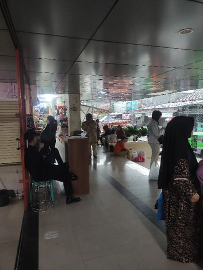People gather in an open market, some standing and others seated. A woman in a black hijab and patterned clothing is prominently positioned near the camera. Other figures include a man in a light-colored shirt and people in more casual attire. The scene suggests a bustling marketplace with a mix of activities and interactions. Bright lighting emphasizes the variety of goods available for purchase, creating a vibrant atmosphere.