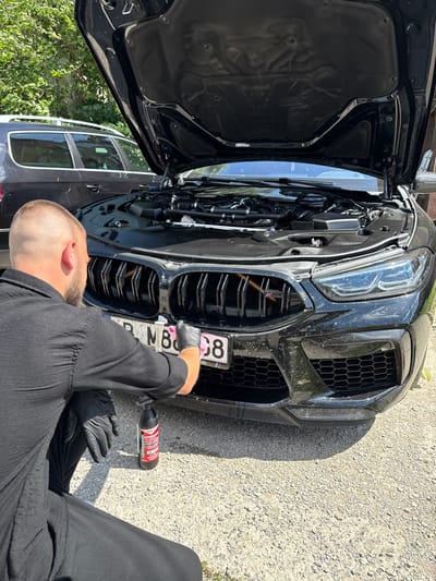 A male individual is observed in a crouched position, wearing a black long-sleeved shirt and black gloves, actively cleaning the front of a black high-performance BMW M-series vehicle. The car's hood is open, exposing components of the engine bay. The vehicle, registered in Targovishte, Bulgaria, as indicated by the "P" on its front license plate (partially visible as "P. MB 8"), displays visible dirt and splatters across its front bumper, large black kidney grilles with an M-series badge, and carbon fiber lower trim. The individual is using a pink microfiber cloth on the grille area, with a black spray bottle labeled "HIPERSONIK" resting on the gravel ground beside his left knee. The scene is outdoors on an unpaved surface of small gray gravel, under bright daylight which casts distinct shadows. In the background, partially visible, is a dark-colored station wagon or SUV, with green foliage from trees and bushes behind it.