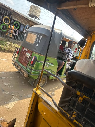 The image depicts a street scene in Umuode, Nigeria, viewed from inside a yellow tricycle. In the foreground, parts of the observer's yellow tricycle are visible, including a rusty yellow metal frame and a black textured seat. A brown animal ear is partially visible in the bottom left.
The central subject is a green TVS Keke Deluxe tricycle (auto-rickshaw) positioned on an unpaved dirt road. The tricycle has a dark grey canopy and features "TVS" on the rear and "DELUXE" and "BOXER" text on its side. Its rear bumper is a metal rack, with brown material (likely mud or dirt) adhered to the lower section. A Nigerian license plate, partially obscured, is visible on the rear. Inside the green tricycle, two individuals are seated: one wearing a red shirt and another wearing a black and white patterned shirt. The leg of a third person, wearing shorts, is also visible.
In the background, on the left, a single-story commercial building with a corrugated metal roof is present. Its facade features vertical metal bars, and several colorful ring-shaped objects (yellow, green, white, pink) are displayed on the exterior, possibly merchandise. Above the building, against a clear sky, an elevated rectangular water tank and utility wires are visible. The ground is a light brown dirt road, scattered with small pieces of white debris. The scene is illuminated by strong daylight.