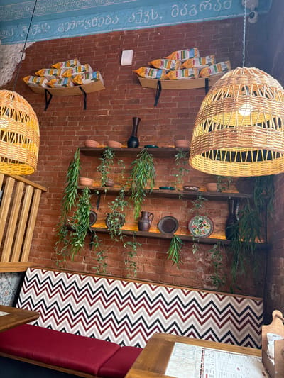 The image depicts an indoor scene, likely a restaurant or cafe. The dominant color is the warm red of the brick wall that forms the backdrop. Several wooden shelves are mounted on the wall, displaying various items. On the top shelf, there are stacks of bagged goods, possibly snacks. The middle and lower shelves hold ceramic bowls and plates, as well as decorative vases and artificial hanging plants.
A large, woven lampshade made of natural material hangs from the ceiling, casting a warm light. To the left, a section of a wooden slatted structure is visible, possibly part of a booth or partition. In the foreground, a banquette seat with a distinctive chevron pattern in red, black, and beige covers the back, and a solid red cushion forms the seat. A wooden table is partially visible in the lower left corner, with a menu laid out on it.
The overall aesthetic suggests a cozy, rustic, and possibly ethnic-inspired decor. The inscription in Georgian script on the blue painted upper part of the wall provides a cultural clue. This establishment could be a Georgian restaurant or a place that embraces Georgian cultural elements. There are no people visible in the image. The lighting is ambient and warm, contributing to the inviting atmosphere.