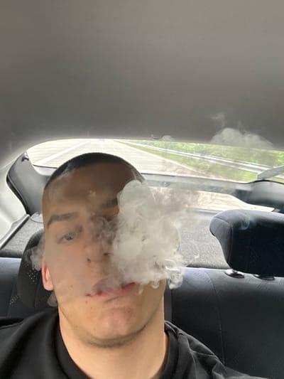 The image shows a person exhaling a large cloud of vapor, presumably from a vape device. The individual has short, dark hair and is wearing a dark-colored shirt. The setting appears to be inside a vehicle, based on the window, seat, and interior panels visible. The background visible through the window suggests an outdoor environment with greenery. The image is not related to the city of Bozduganovo, Bulgaria.